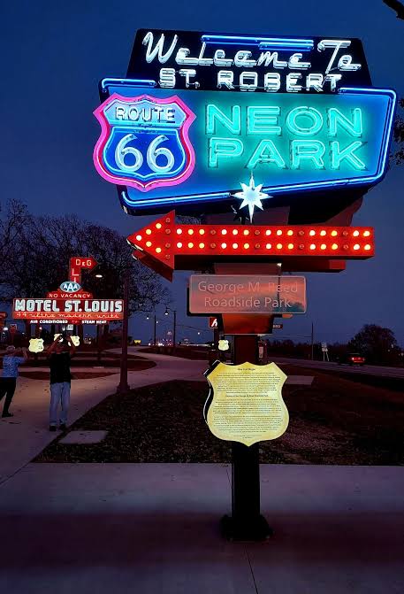 Neon signs at dusk: a large “Welcome to St. Robert Route 66 Neon Park” sign with an arrow, and a lit “Motel St. Louis” sign in the background, with people standing nearby on the sidewalk.