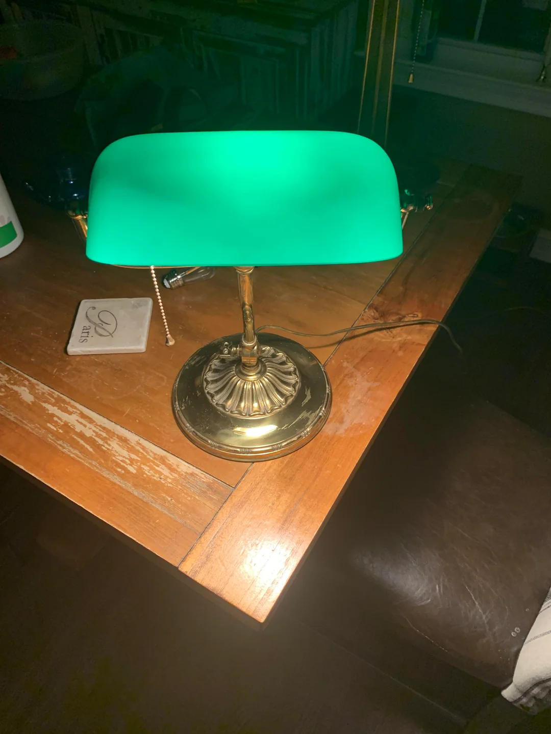 A brass desk lamp with a green glass shade sits on a wooden table, turned on and illuminating the area. A coaster and some papers are beside it, and a leather chair is partially visible.