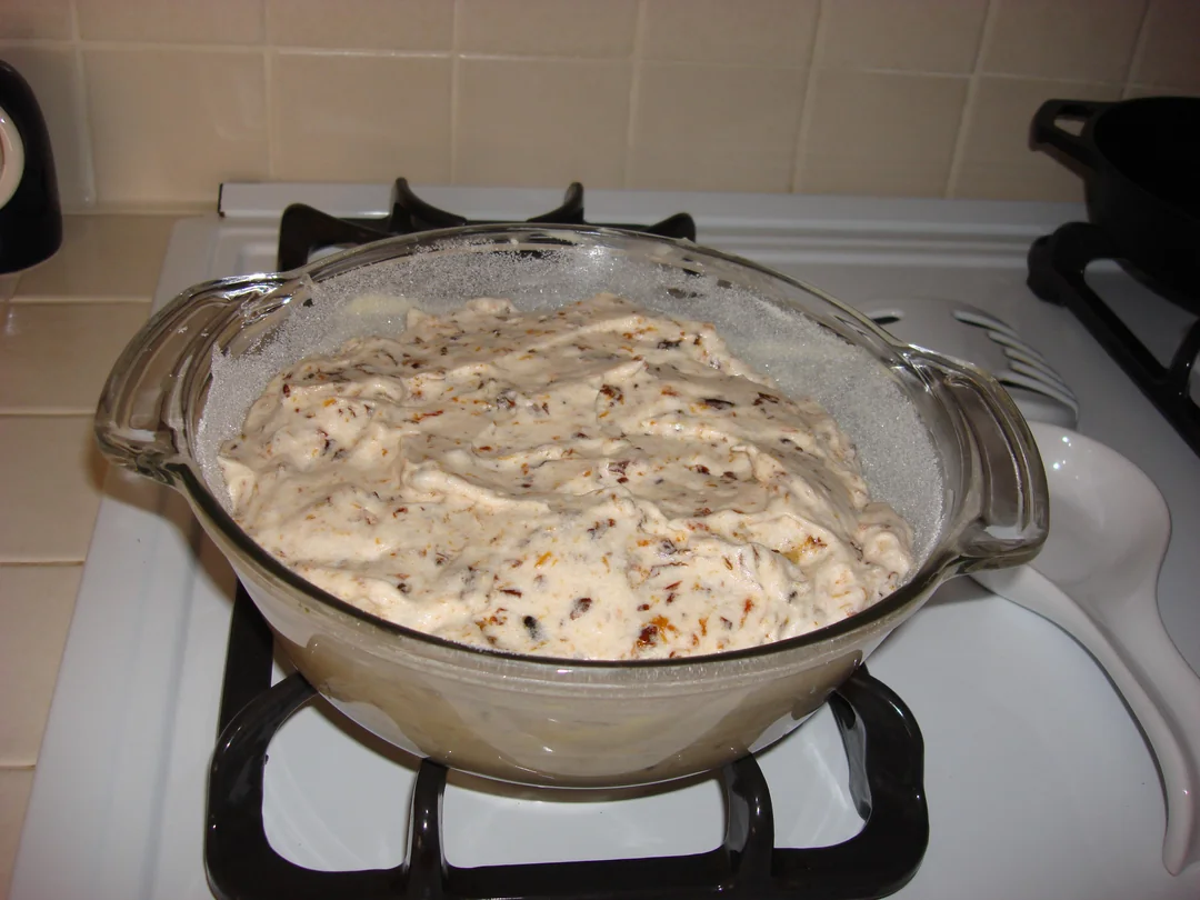 A glass dish filled with uncooked batter or dough with visible specks, sitting on a stove top in a kitchen with beige tiled backsplash.