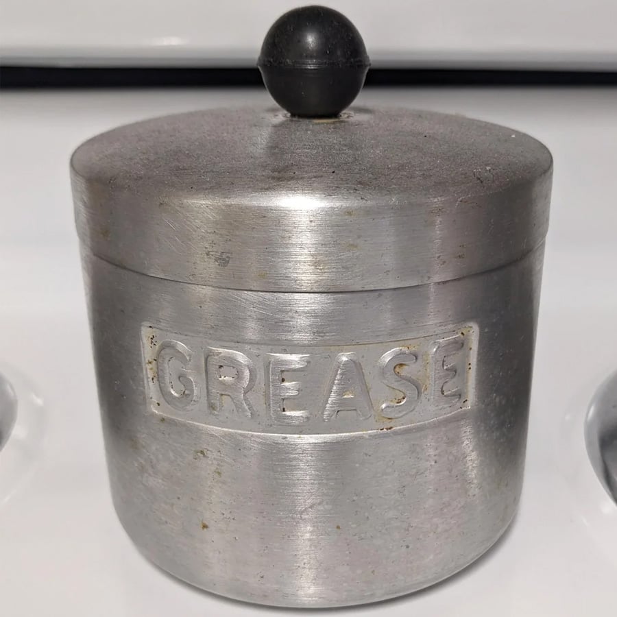 A metal container with a lid and knob handle, labeled “GREASE” on the front, sits on a white stovetop.