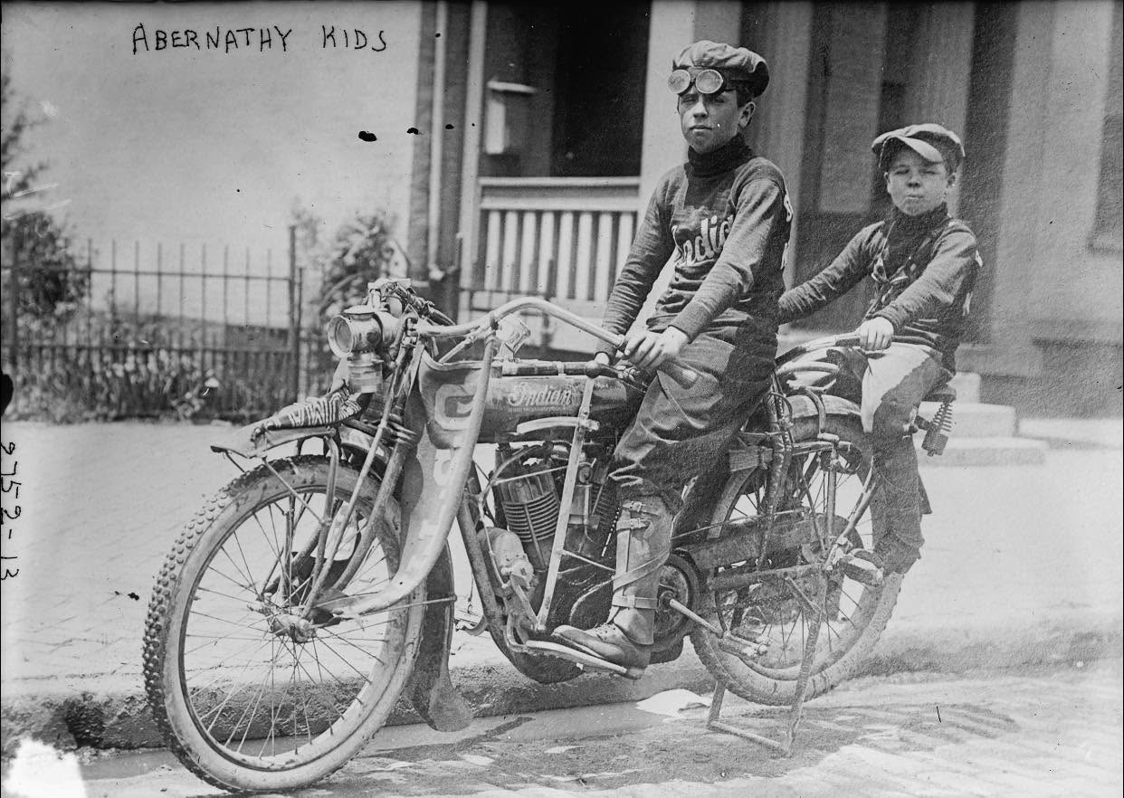 Two boys sit on an old motorcycle in front of a house, wearing goggles and hats. The words “ABERNATHY KIDS” are written above them. The photo is black and white and appears to be historical.