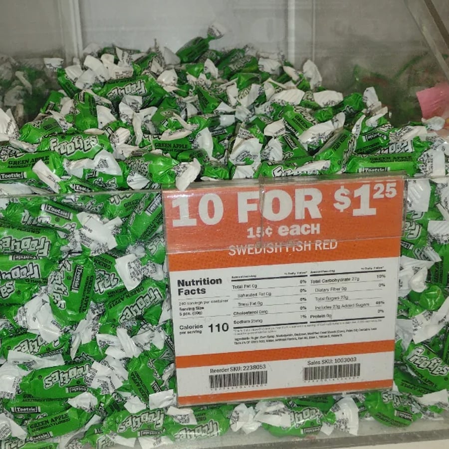 A bin filled with green and white Sour Apple Tootsie Rolls, with a sign reading “10 for $1.25, 15¢ each” and nutrition facts below the price.