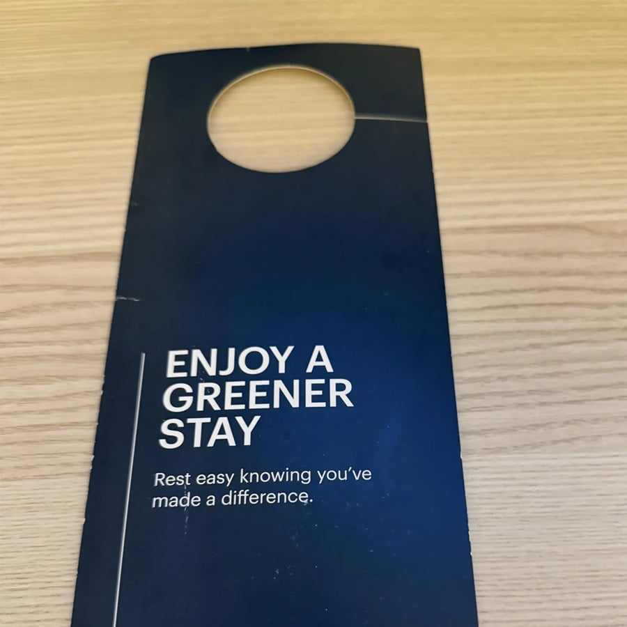 A navy blue door hanger on a wood surface reads, “ENJOY A GREENER STAY. Rest easy knowing you’ve made a difference.”