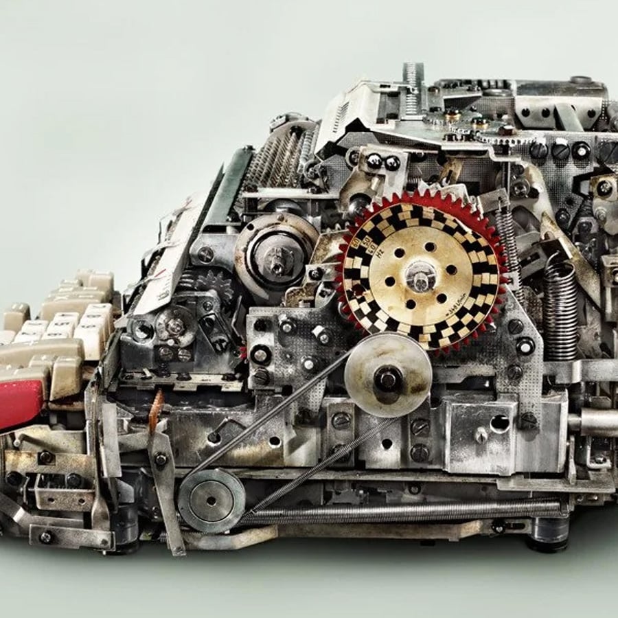 A close-up of a complex mechanical device made from various metal gears, springs, and levers, resembling a typewriter or old machine, set against a plain light background.