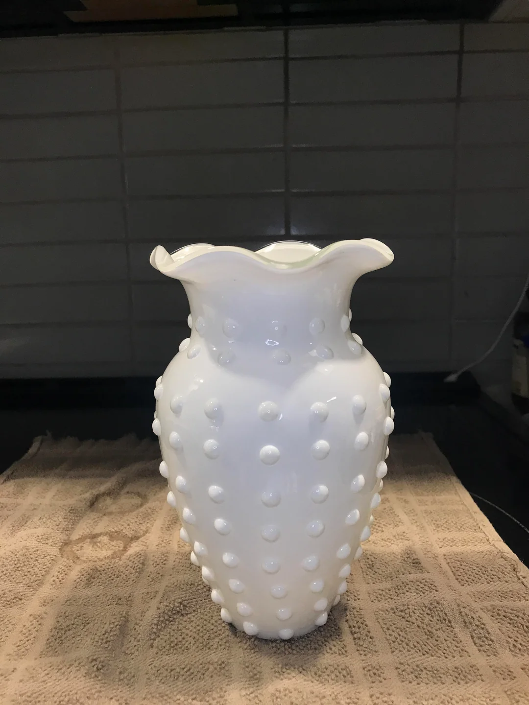A white hobnail-patterned glass vase with a ruffled rim sits on a beige textured towel, against a background of white tiled wall.