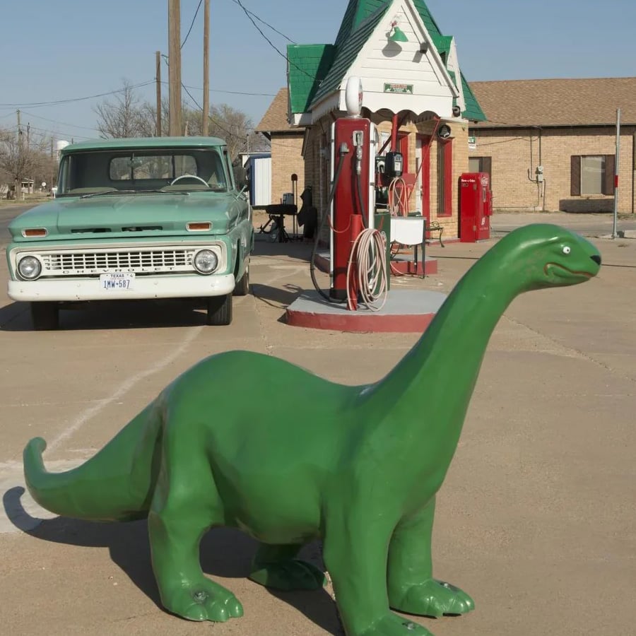 A green dinosaur statue stands in front of a vintage green truck and an old-fashioned gas station with a red pump on a sunny day.