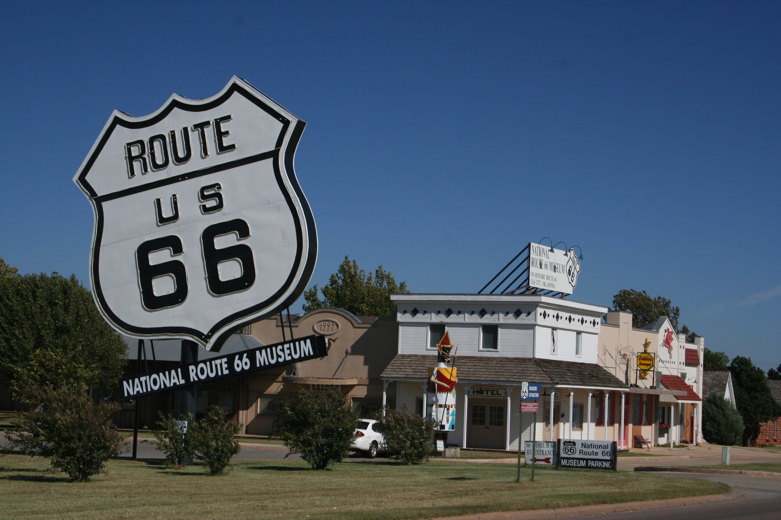 Large Route 66 sign in front of the National Route 66 Museum, with vintage-style buildings and a clear blue sky in the background.