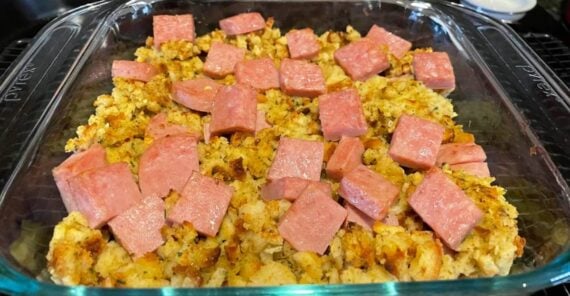 A glass baking dish filled with a casserole of cubed pink meat and seasoned bread stuffing, sitting on a cooling rack with a brick backsplash in the background.