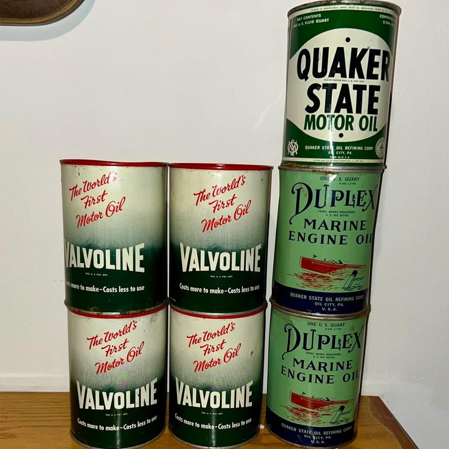 Six vintage Valvoline and two vintage Quaker State and Duplex oil cans are stacked against a wall. The cans feature retro designs, bold typography, and are arranged in two columns.
