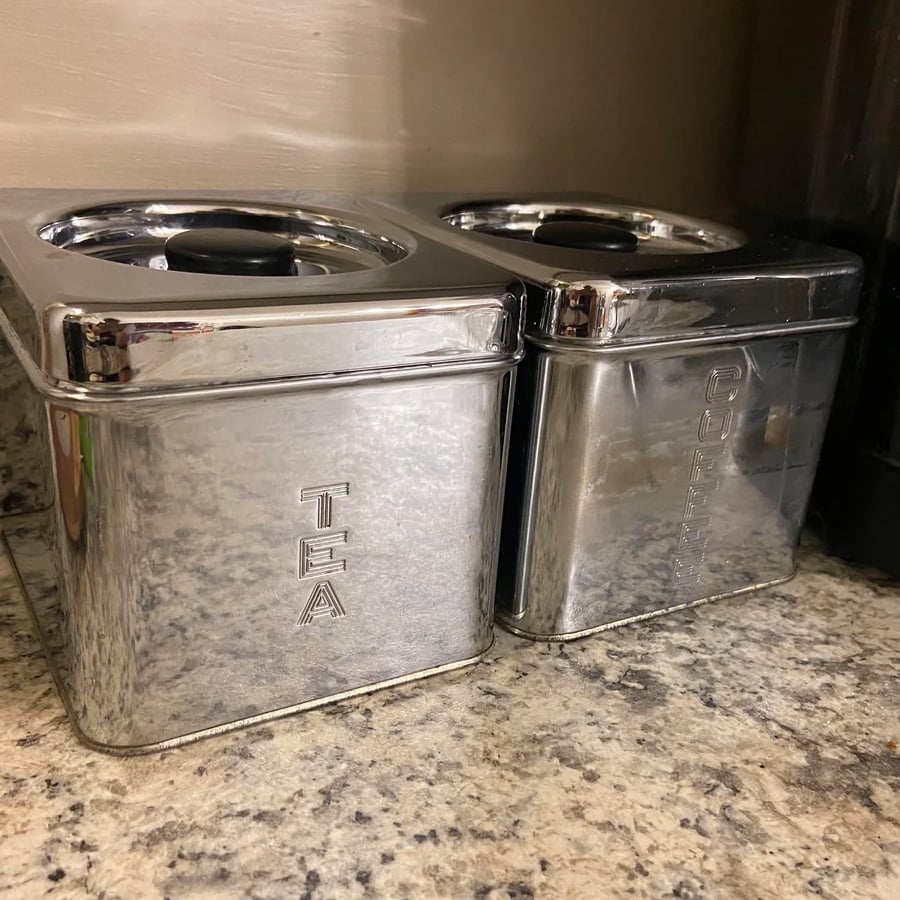 Two shiny metal containers sit on a granite countertop. One is labeled “TEA” and the other is labeled “COFFEE,” both with vertically engraved letters on their front sides.