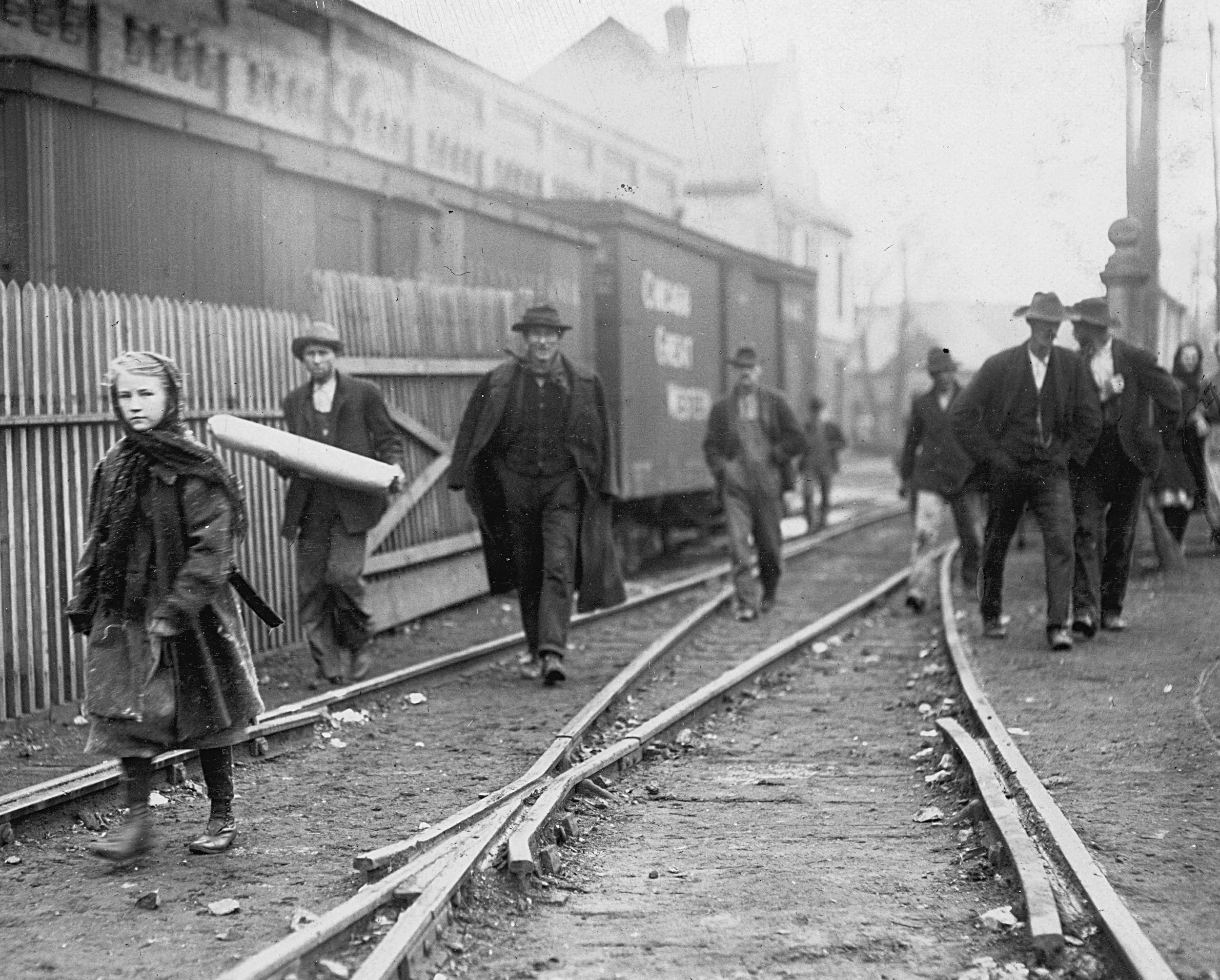 Black-and-white photo showing people walking along train tracks. A young girl with a shawl walks in the foreground, while men in hats and coats walk behind her. A train car and wooden fence are visible. The setting appears historical.