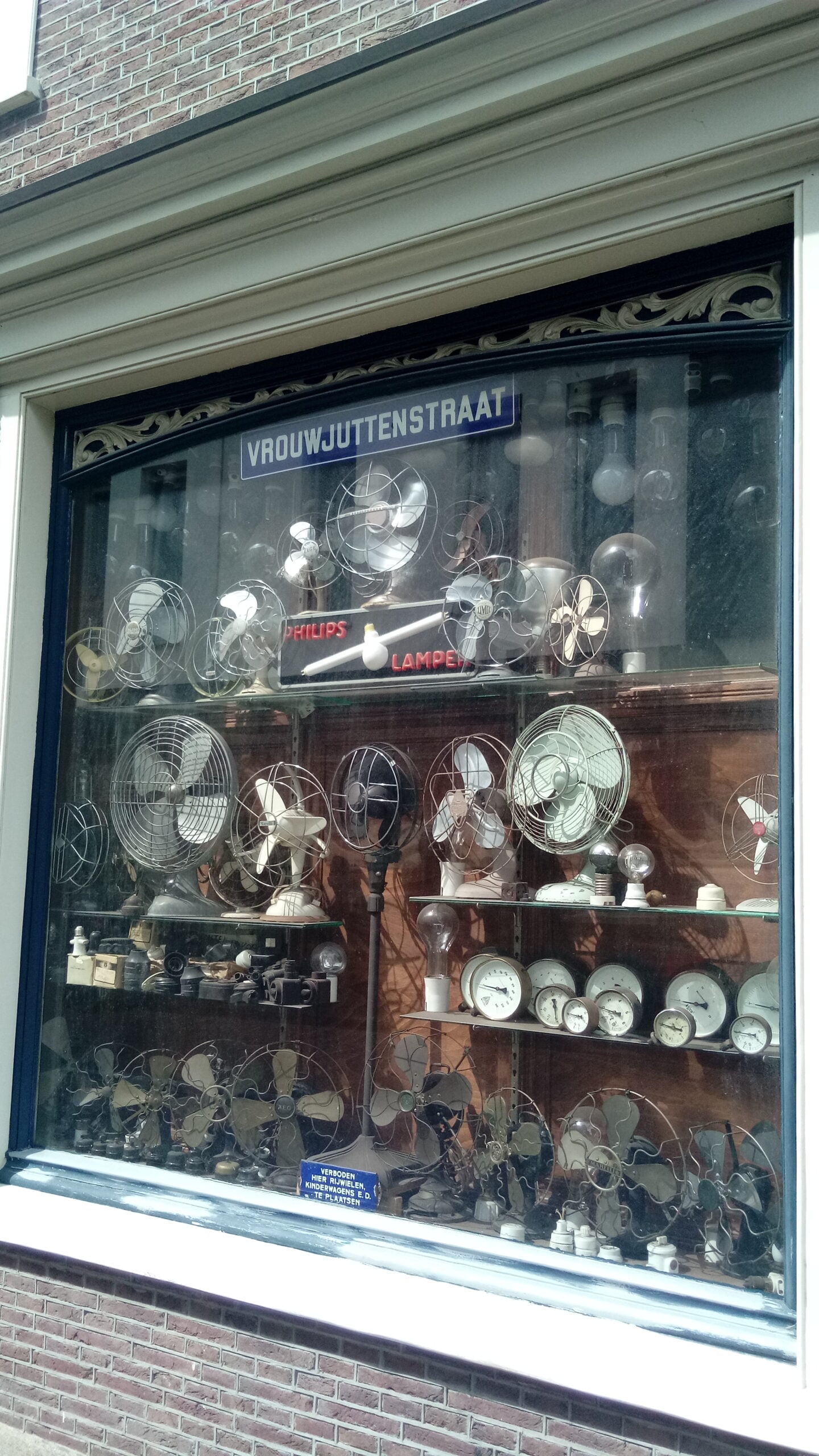 A shop window on Vrouwjuttenstraat displays vintage electric fans, clocks, and light bulbs. The reflection of buildings is visible in the glass.