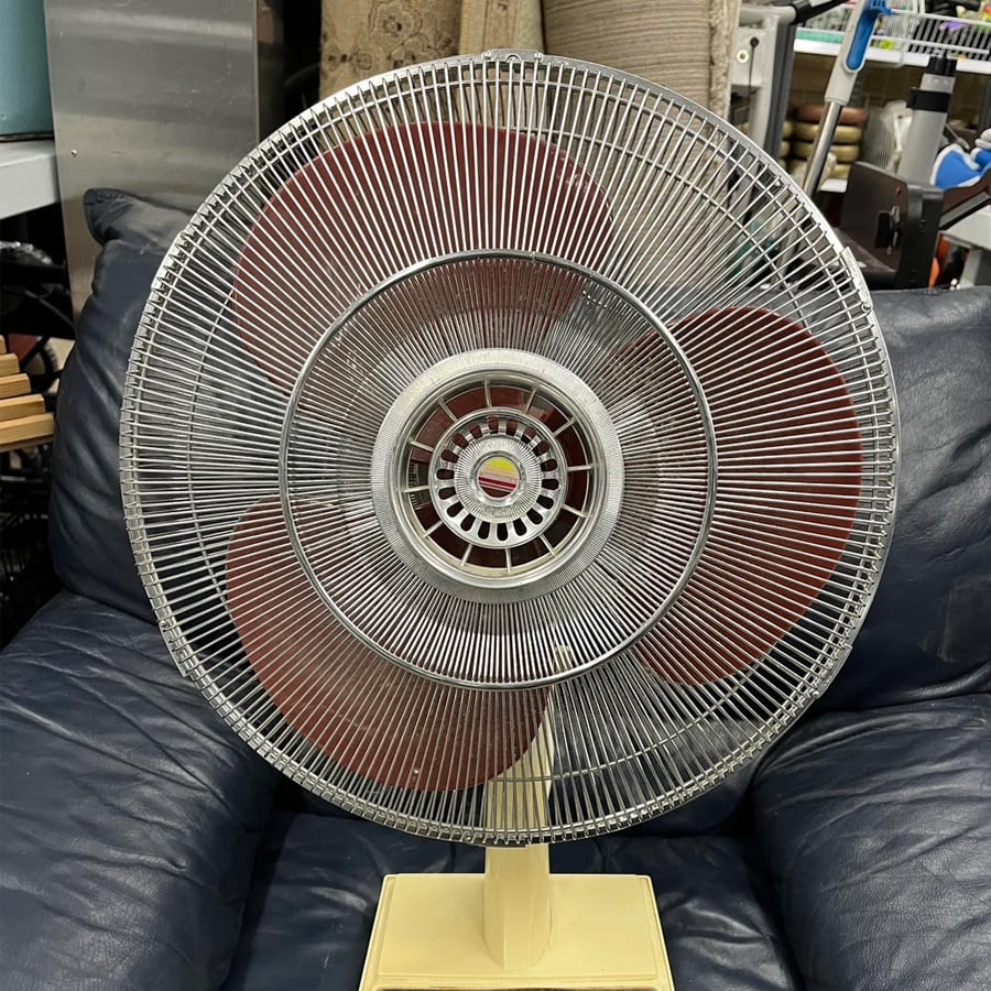 A vintage electric fan with red blades and a metal grill sits on a dark blue cushioned chair in a cluttered room with shelves and various items in the background.