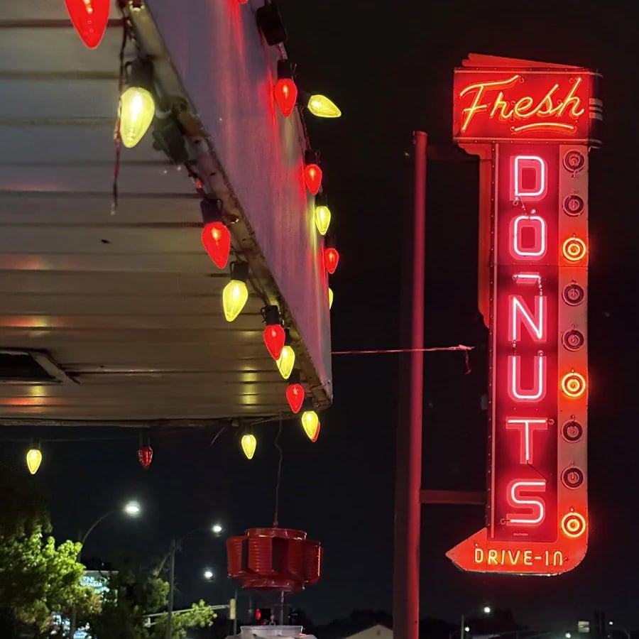 Retro neon sign reads “Fresh DONUTS DRIVE-IN” at night, next to a building decorated with red and yellow string lights. Trees and streetlights are visible in the background.