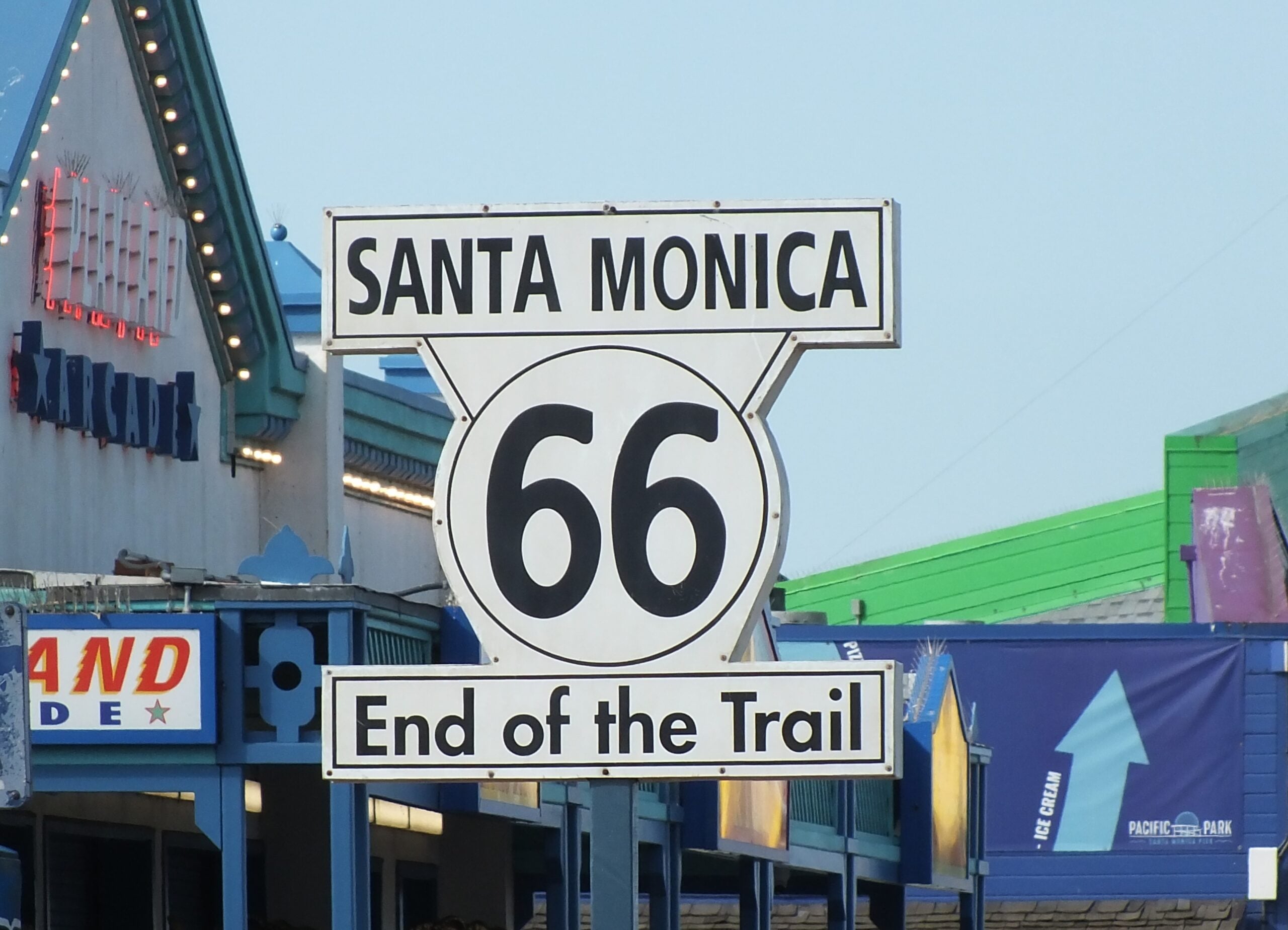 Route 66 “Santa Monica End of the Trail” sign, located at the Santa Monica Pier, with colorful buildings and signs in the background.
