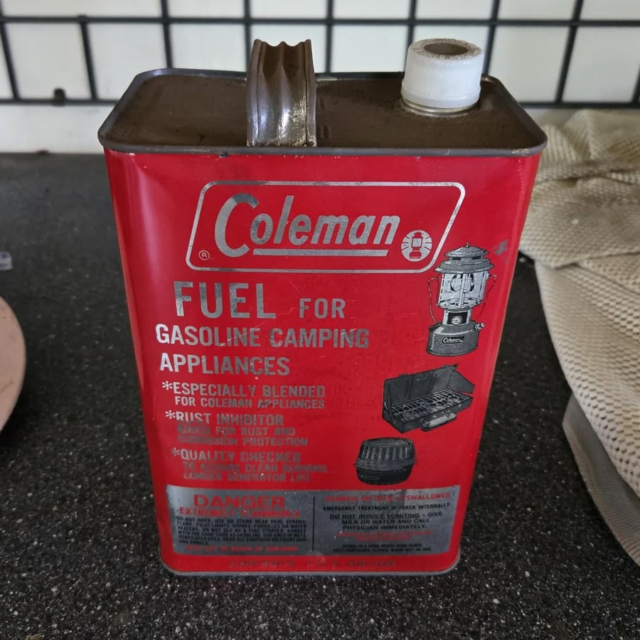 A red metal Coleman can labeled “Fuel for Gasoline Camping Appliances” sits on a counter. The can shows product details and illustrations of camping stoves and lanterns.