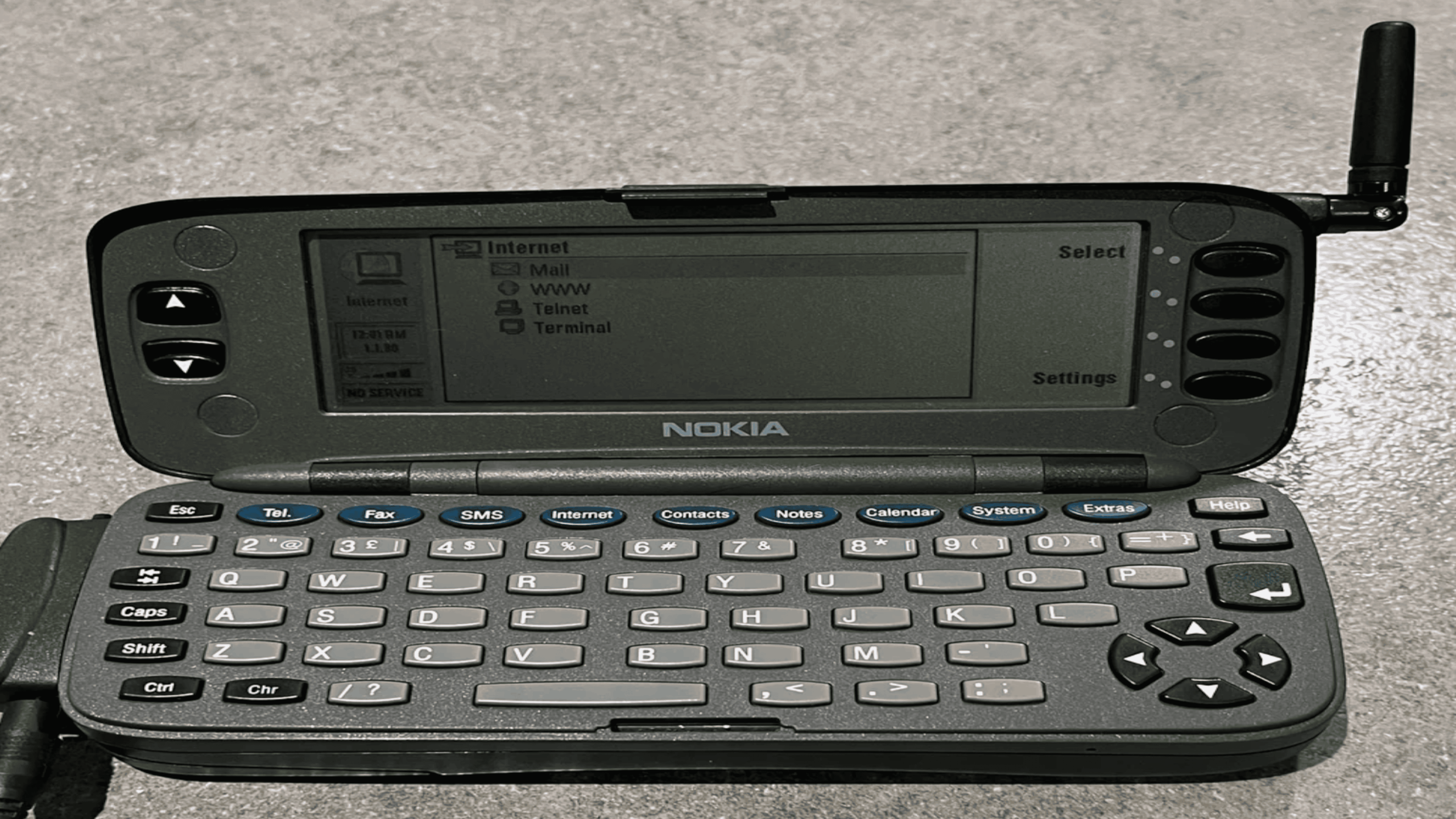 A vintage Nokia Communicator phone with a full QWERTY keyboard, screen displaying internet options, and an extended antenna, sits open on a gray surface.
