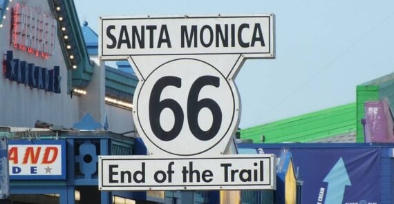 Close-up of a Route 66 “End of the Trail” sign in Santa Monica, set against a blue sky with colorful buildings and signs in the background.