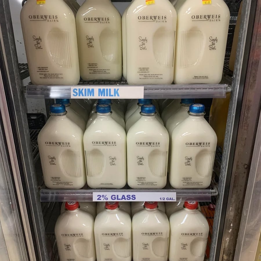 Glass bottles of Oberweis Dairy skim milk and 2% milk are displayed on a refrigerated store shelf. The bottles are labeled “Simply the Best.” A sign labels the section for “SKIM MILK” and “2% GLASS” options.