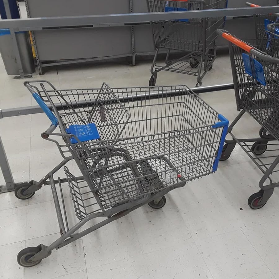 A shopping cart with a bent frame and tilted basket sits beside other normal carts on a store’s white tile floor.
