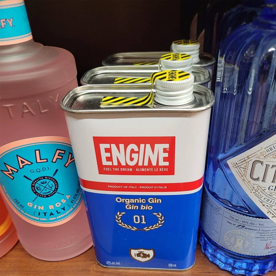 A rectangular blue and white can labeled “ENGINE Organic Gin” sits on a shelf between pink and blue bottles of gin. The can has a metal cap with a yellow and black striped pull tab.