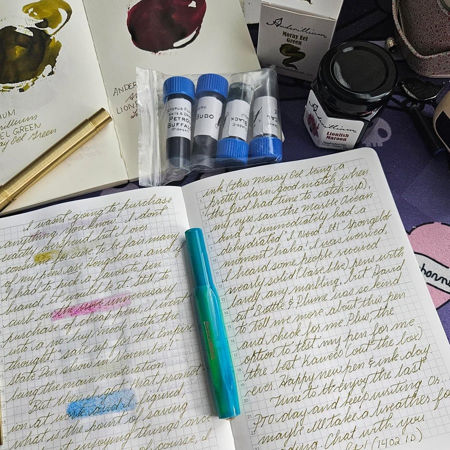 Open notebook with handwritten text in green ink, blue pen on top, ink samples, a gold pen, ink bottles, and another notebook with ink swatches are arranged around it on a table.