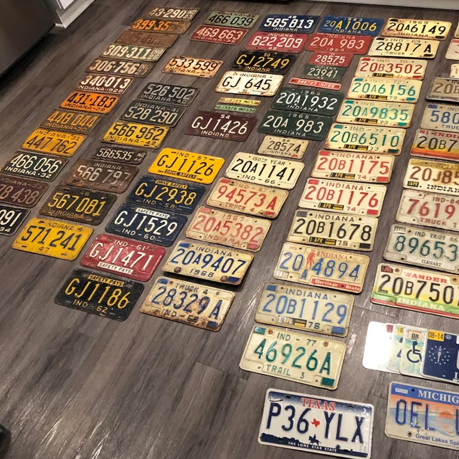 Dozens of vintage license plates from various states are laid out in rows on a wooden floor, displaying a range of colors, numbers, and styles.