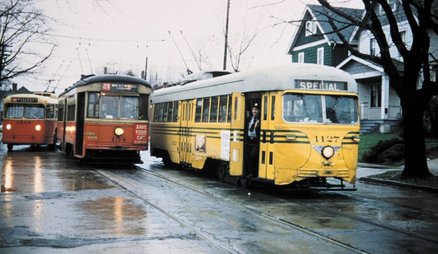 Three vintage streetcars in red and yellow colors travel down a wet street lined with houses and bare trees on a gloomy day. A person stands by the door of the yellow streetcar labeled “SPECIAL.”