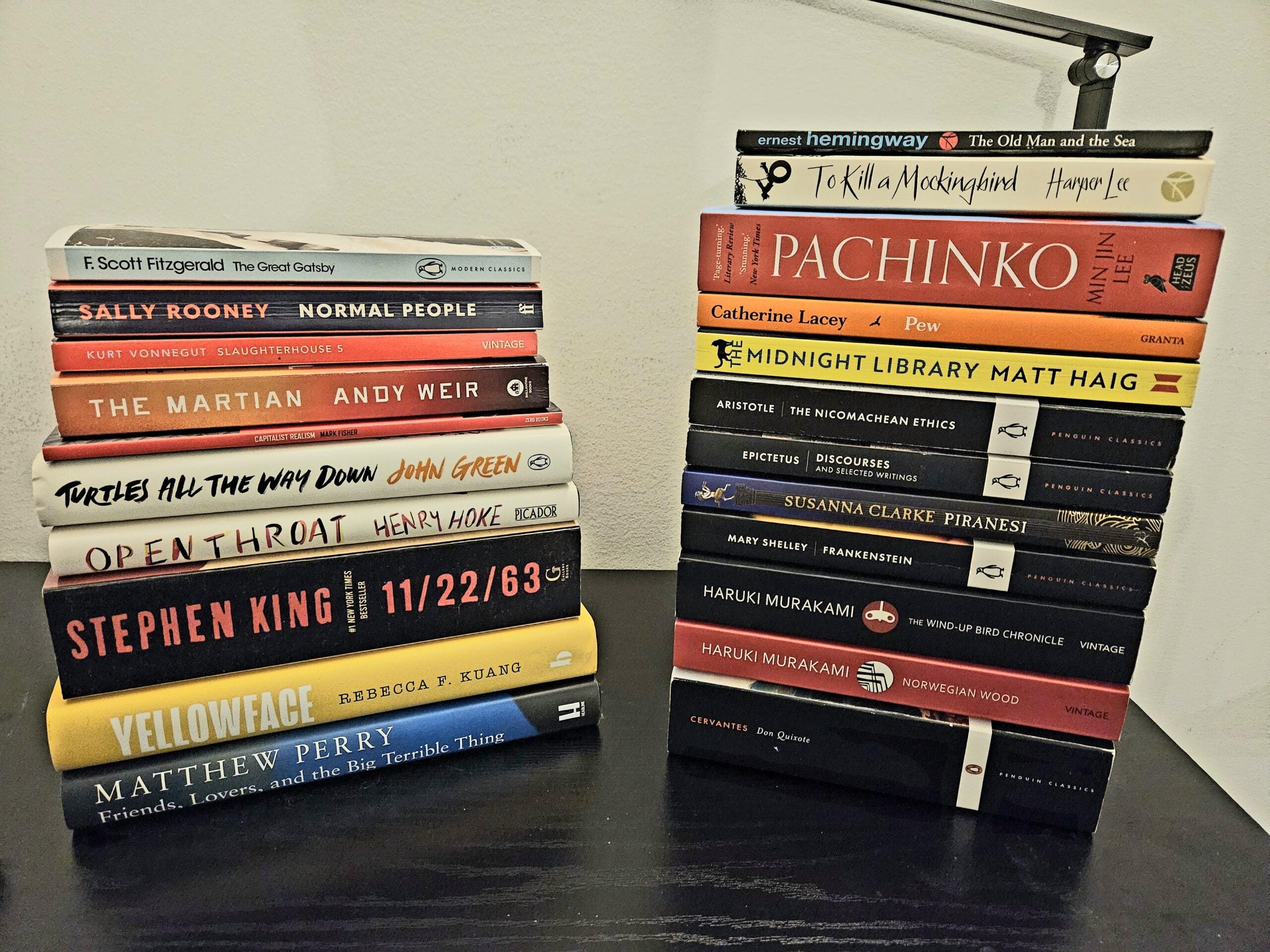 Two stacks of books on a white surface. The left stack includes titles like “The Great Gatsby” and “11/22/63.” The right stack includes “The Old Man and the Sea,” “Pachinko,” and several Haruki Murakami novels.