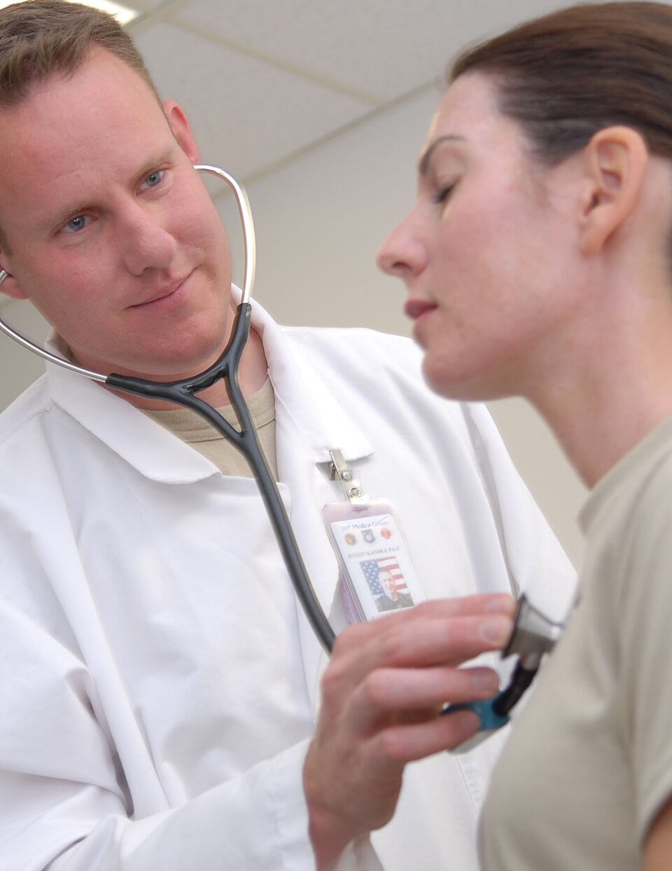 A doctor wearing a white coat uses a stethoscope to listen to a woman’s chest during a medical examination in a clinic setting.