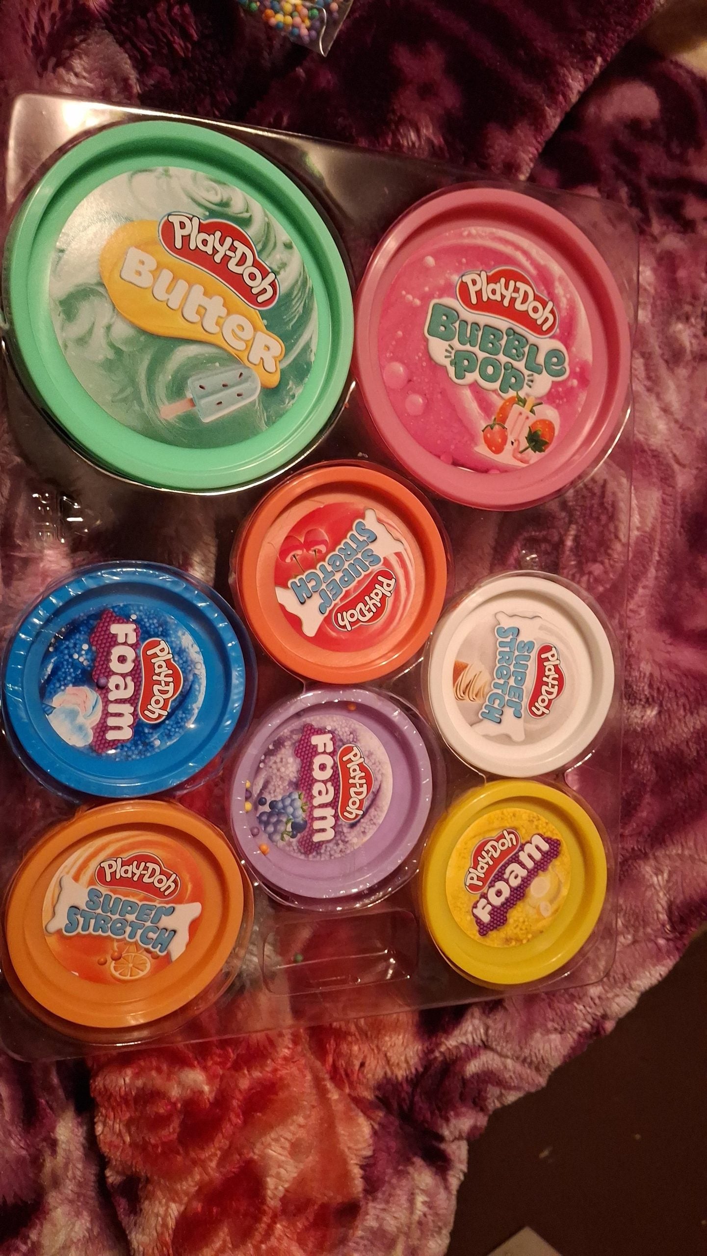A set of nine colorful Play-Doh containers labeled with names like Butter, Bubble Pop, Crystal Crunch, HydroGlitz, Super Stretch, and Foam, arranged on a clear tray atop a purple patterned fabric.