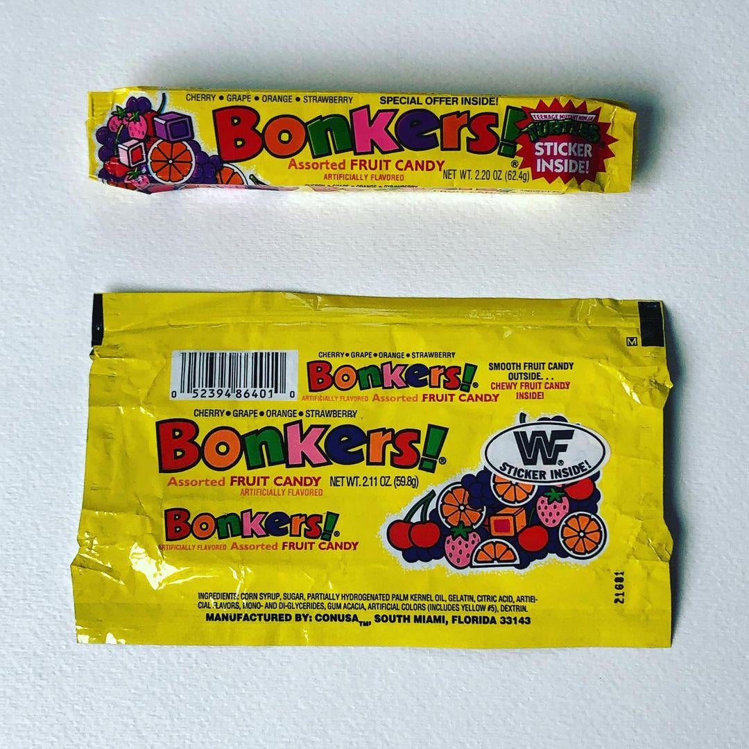 Two yellow Bonkers! candy wrappers with fruit illustrations and colorful text. One wrapper is intact; the other is flattened with a barcode and manufacturing info visible. Both advertise a sticker included inside.