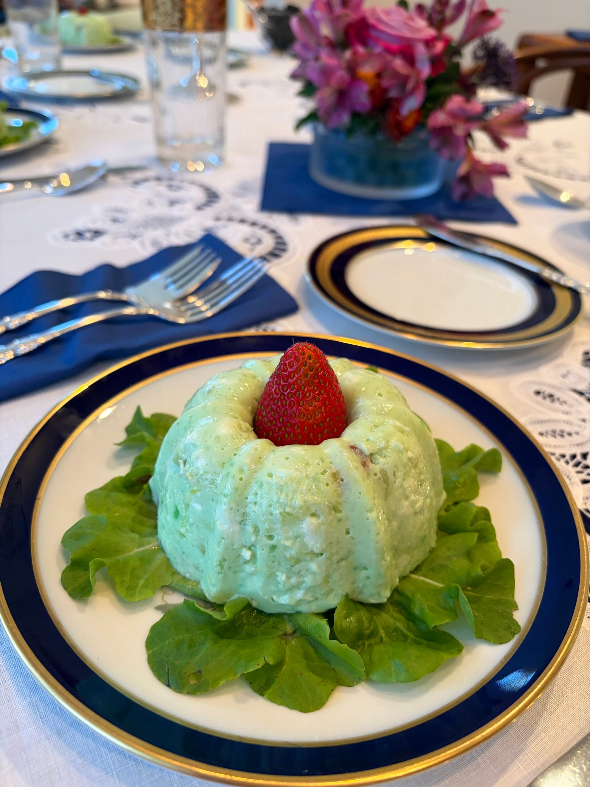 A molded green salad sits on lettuce, topped with a strawberry, on a blue-rimmed plate. The table is elegantly set with silverware, a glass, and flowers in the background.