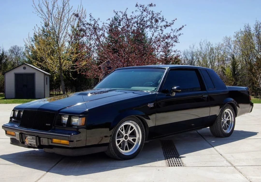 A black two-door coupe with tinted windows and silver wheels is parked on a concrete driveway, with trees and a small shed in the background under a clear sky.