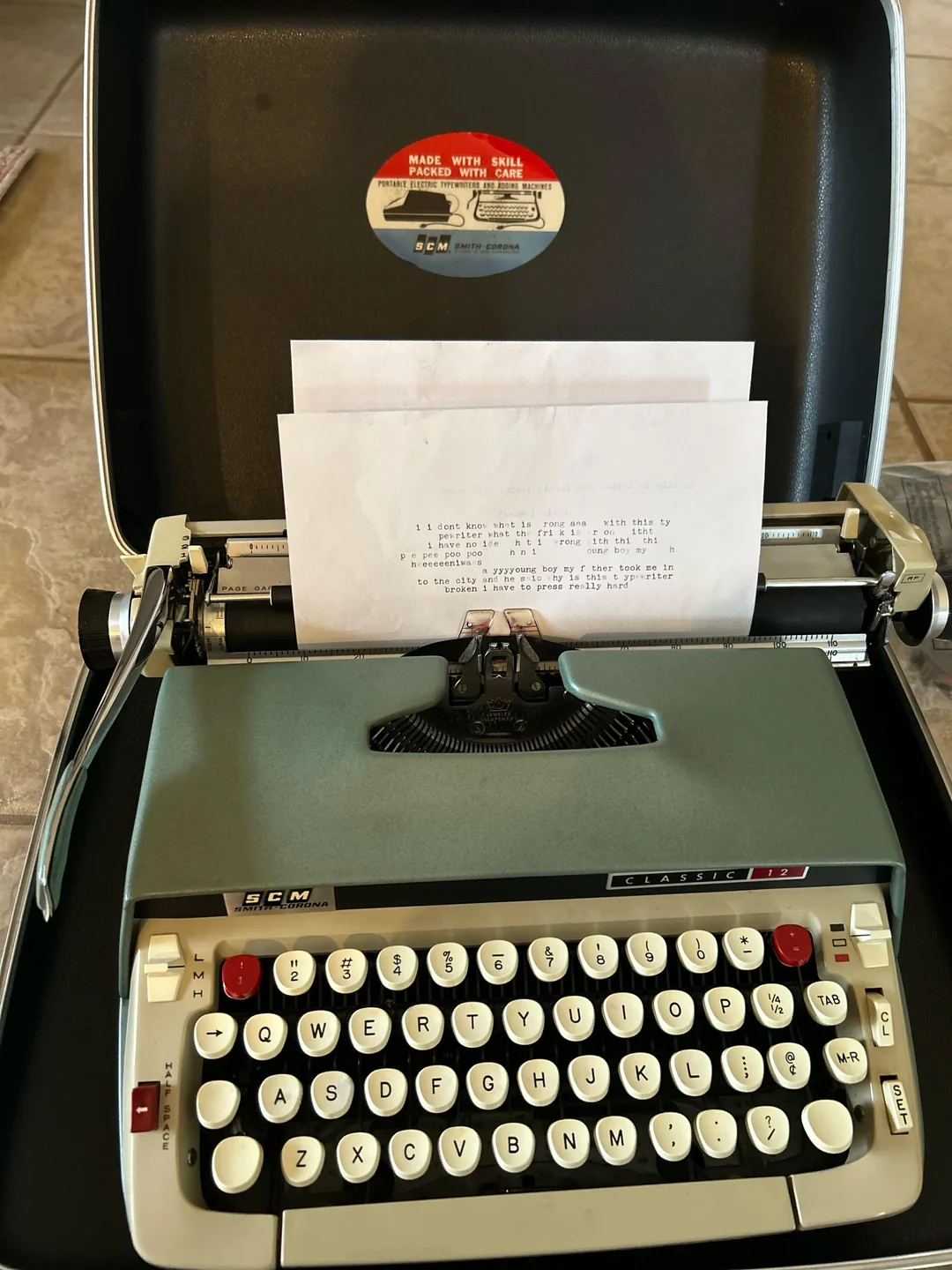 A vintage SCM typewriter with a typed sheet of paper inserted, displayed on a tiled surface. The typewriter is open in its carrying case.