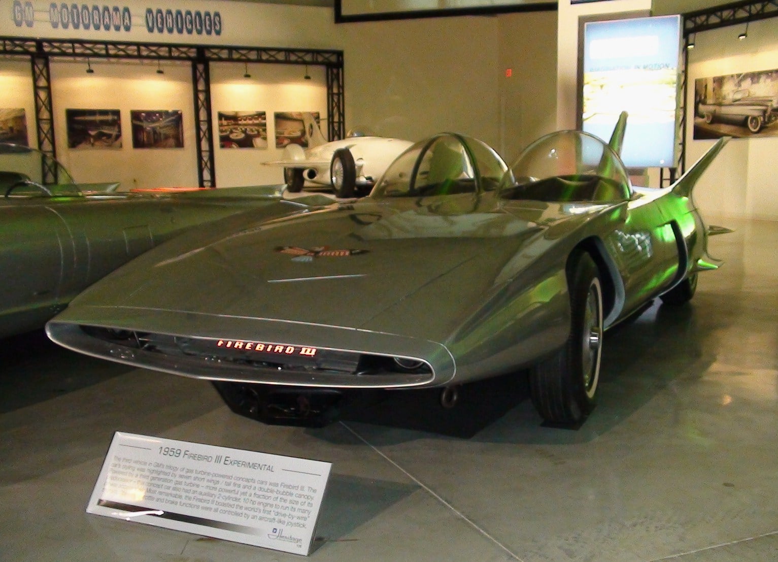 A futuristic silver 1959 Pontiac Firebird III concept car with bubble canopies and finned tail is displayed in a museum setting, with an informational sign in front of it.