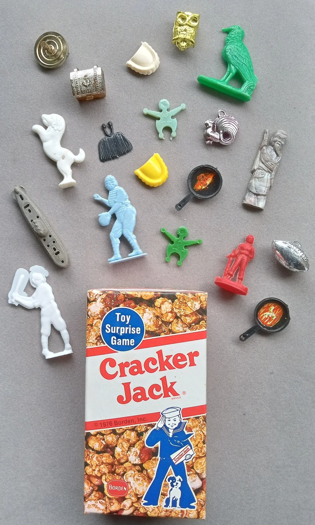 A vintage Cracker Jack box is surrounded by various small plastic and metal toy prizes, including animals, figures, a flute, pans, and other novelty shapes, all arranged on a gray background.