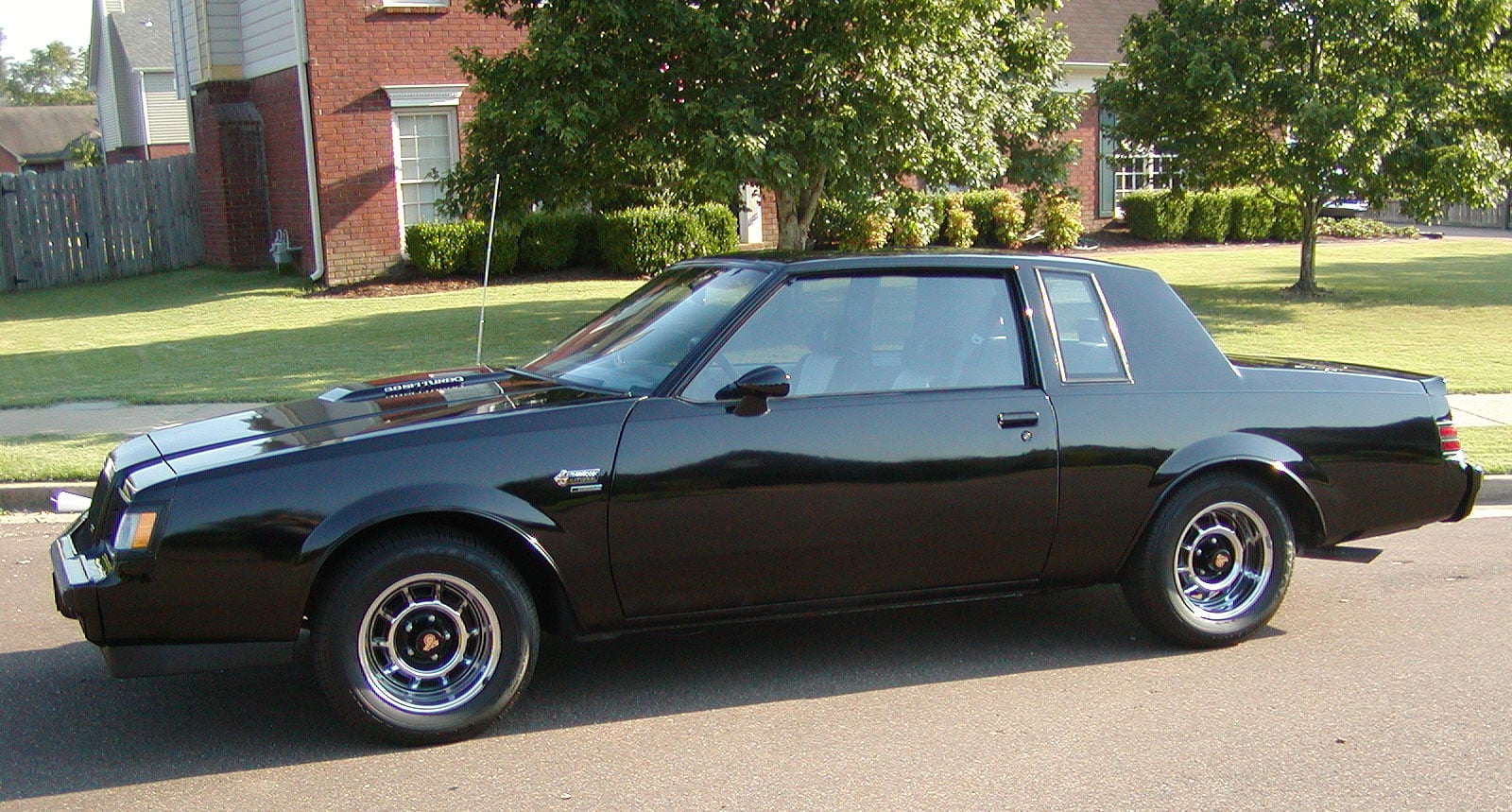 A shiny black classic car is parked on a suburban street with green grass, trees, and brick houses in the background.