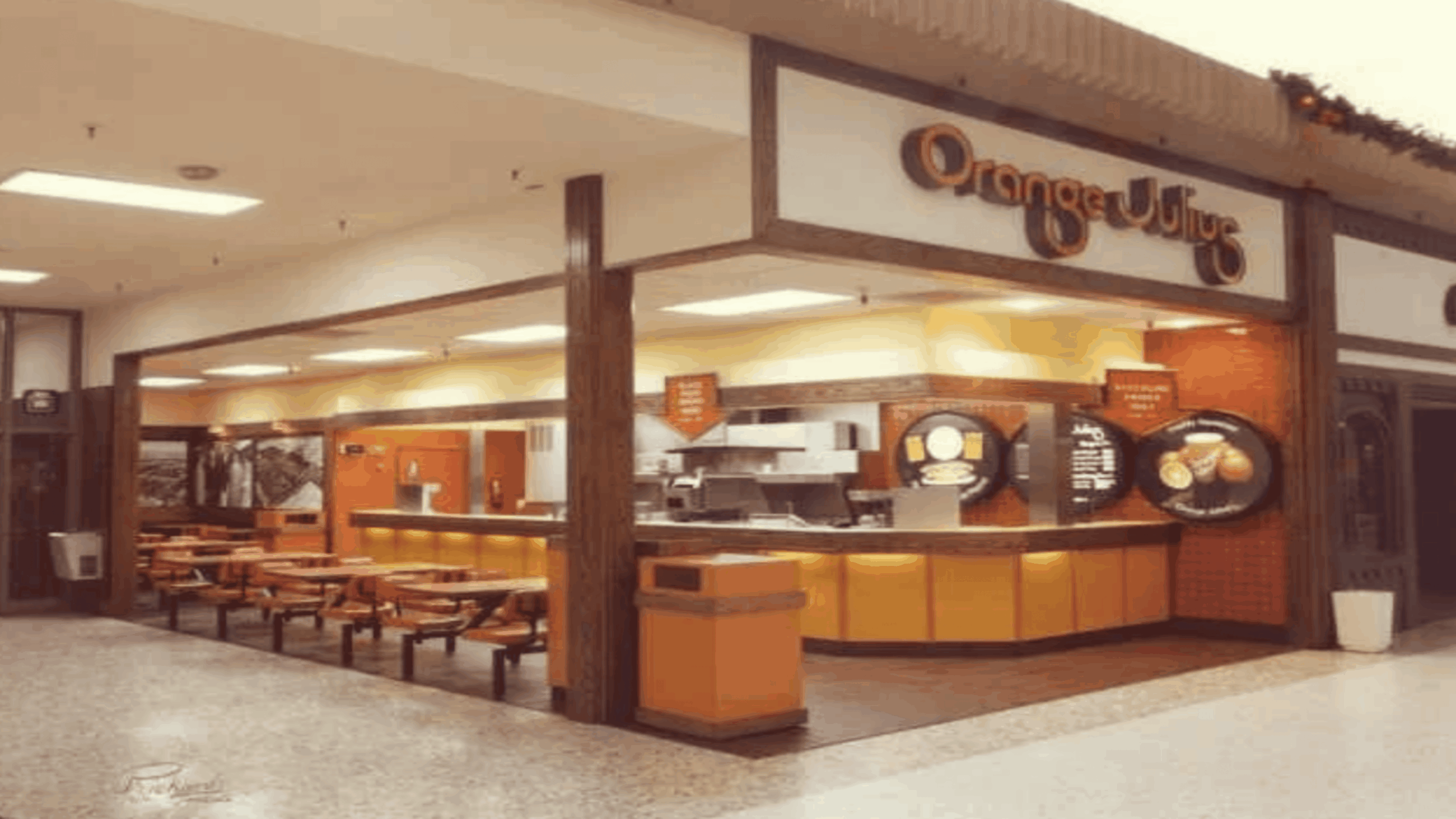 A retro-style Orange Julius mall kiosk with orange and yellow seating, a counter for ordering, menu boards, and a circular Orange Julius sign above the store entrance.