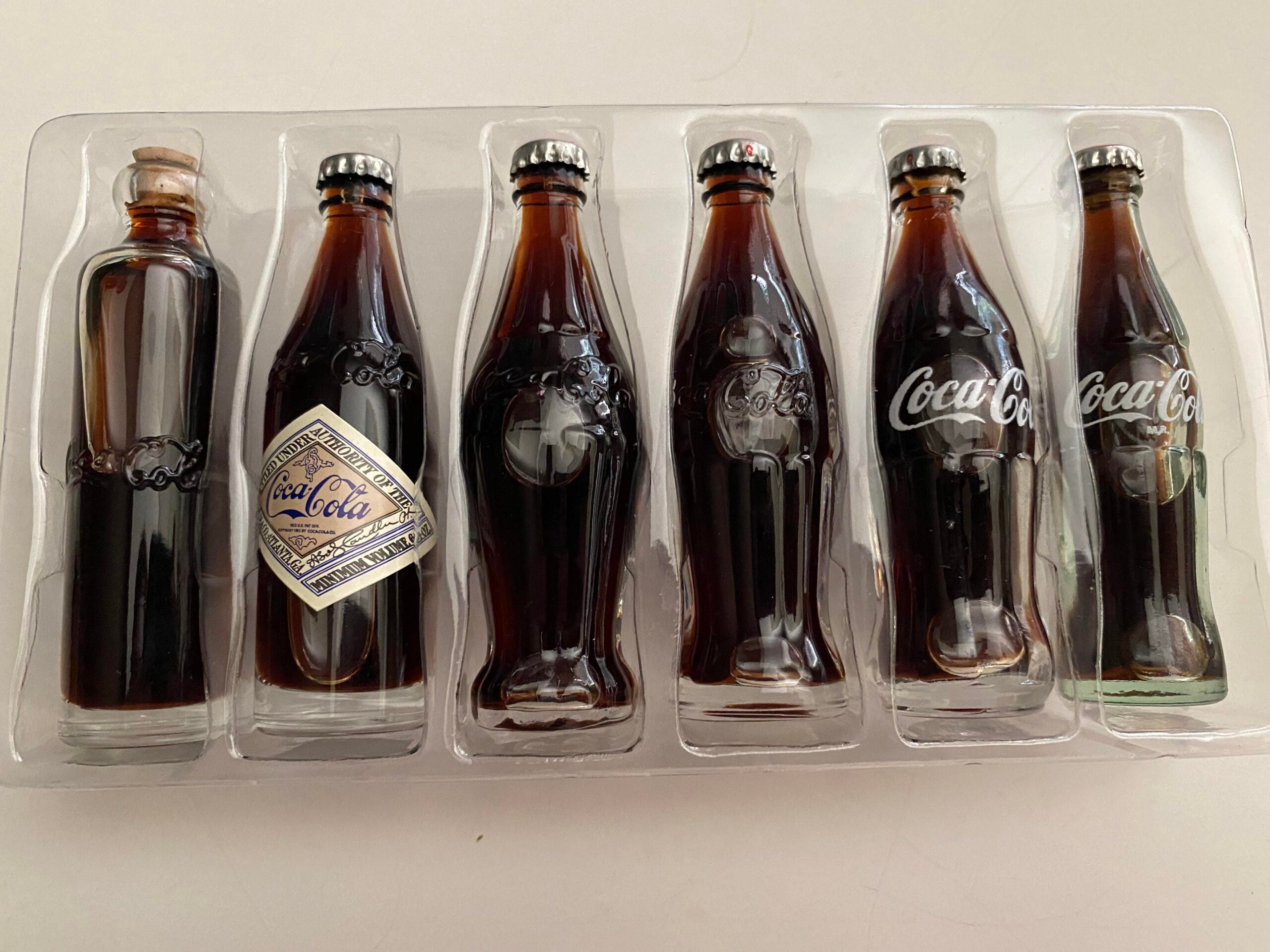Six Coca-Cola glass bottles in a plastic display case, each with a different label or cap design, showing the evolution of Coca-Cola branding over time. Bottles contain dark liquid and are arranged side by side.