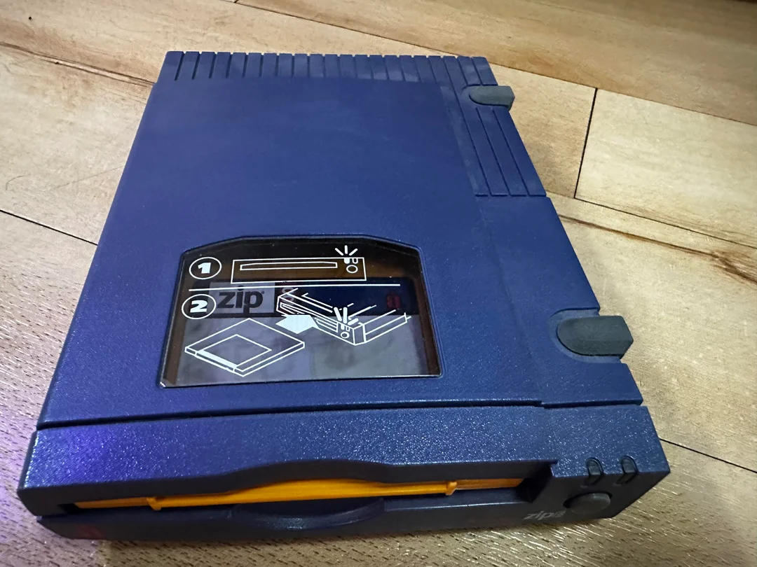 A blue external Zip drive rests on a wooden surface, featuring a clear window with illustrated instructions for inserting a Zip disk.