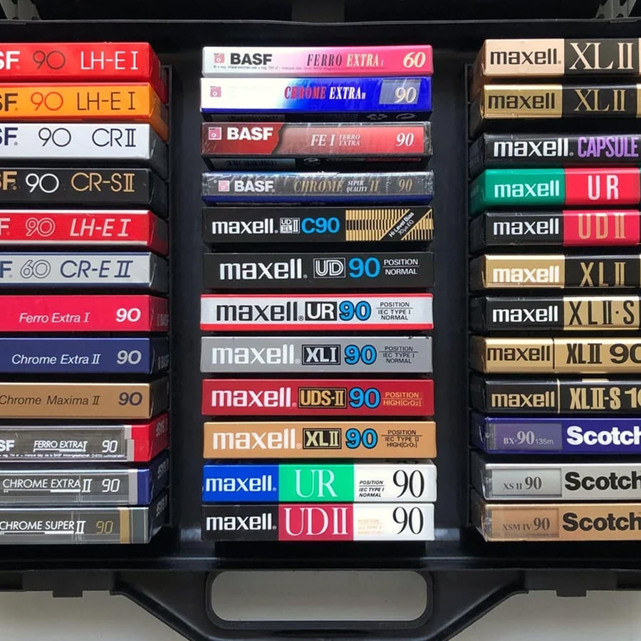 A collection of vintage audio cassette tapes from various brands, stacked neatly in rows within a black storage case. Labels display different models and capacities.