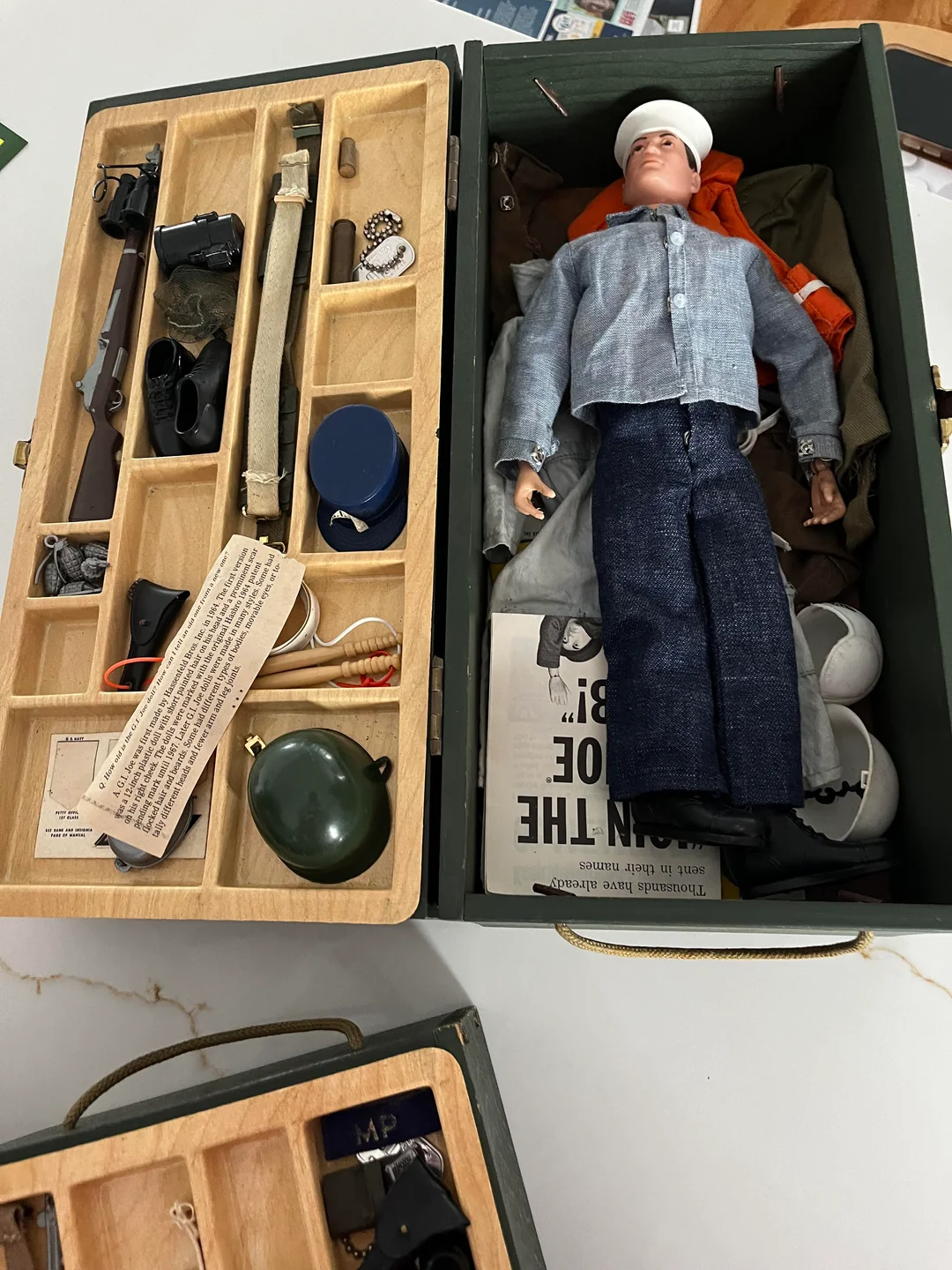 A vintage military action figure in a case, surrounded by miniature accessories like guns, helmet, canteen, patches, and clothing, all neatly organized in compartments.
