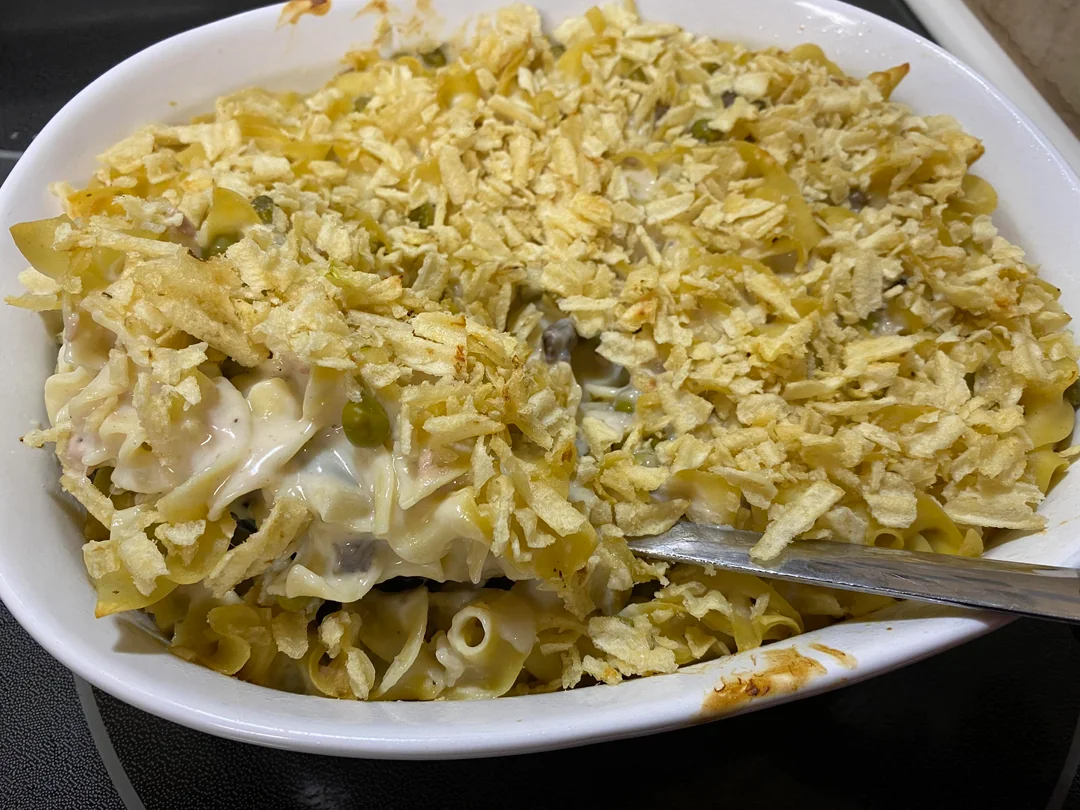 A casserole topped with crushed potato chips, filled with noodles, peas, mushrooms, and a creamy sauce. A serving has been scooped out with a metal spoon.
