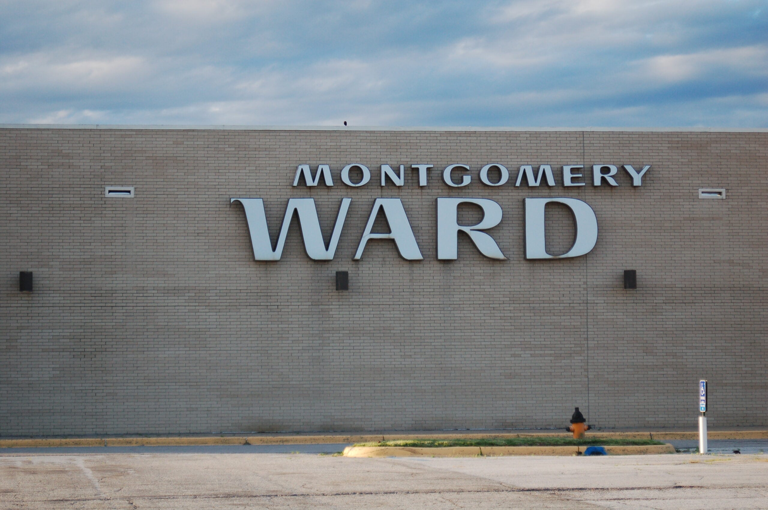 Large “Montgomery Ward” sign on a plain beige brick building, with a mostly empty parking lot and cloudy sky above.