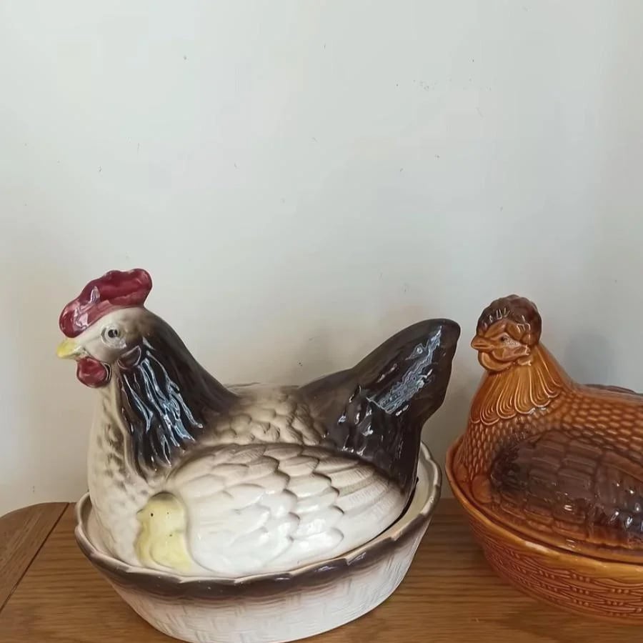 Two ceramic hen-shaped containers sit on a wooden surface against a plain light-colored wall. The larger container is white and black, while the smaller one is brown.