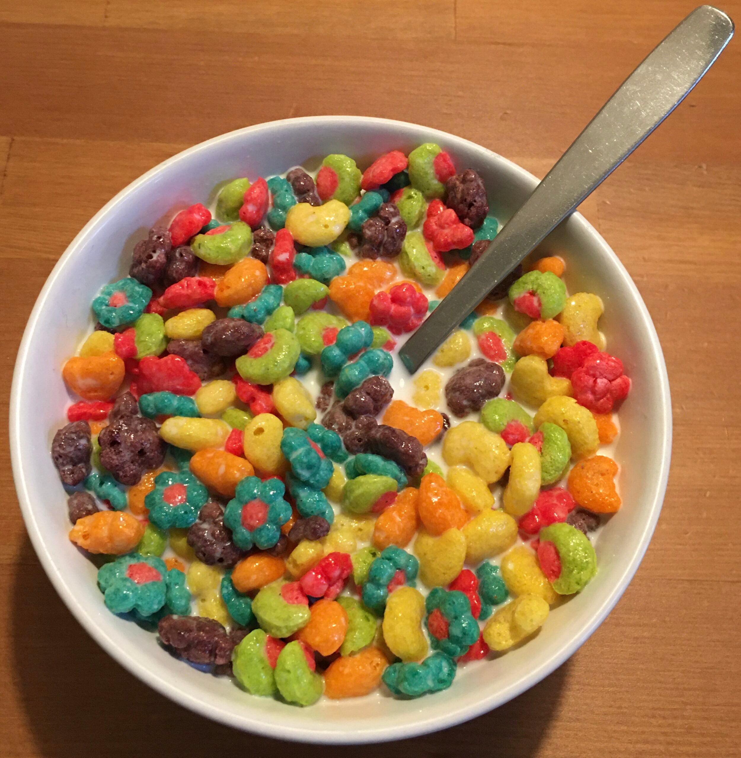 A white bowl filled with colorful, fruit-shaped cereal pieces and milk, with a metal spoon resting inside, sits on a light brown wooden surface.
