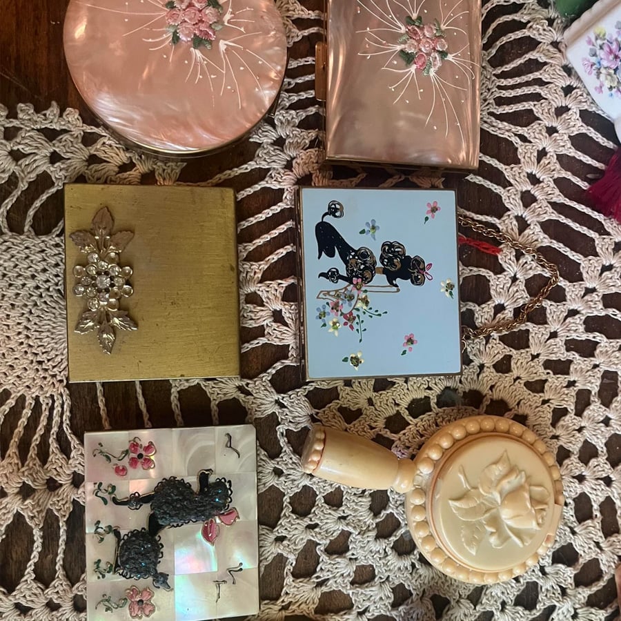 Vintage compacts and a perfume bottle with floral and poodle designs are arranged on a crocheted tablecloth, showcasing intricate details and pastel colors.
