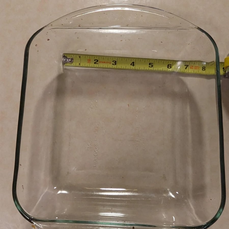 A glass baking dish with a tape measure across the top shows the dish is about 8 inches wide. The dish is clear and square-shaped, resting on a light-colored surface.