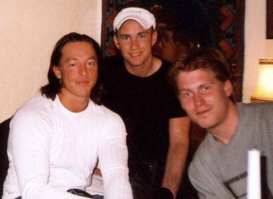 Three young men are sitting together indoors, smiling at the camera. One wears a white shirt, one a black shirt and white cap, and another a gray shirt. A lamp and artwork are visible in the background.